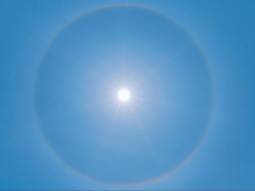 A shining sun in a bright blue sky with a faint rainbow corona.