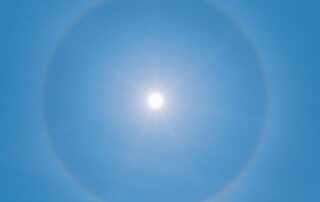 A shining sun in a bright blue sky with a faint rainbow corona.